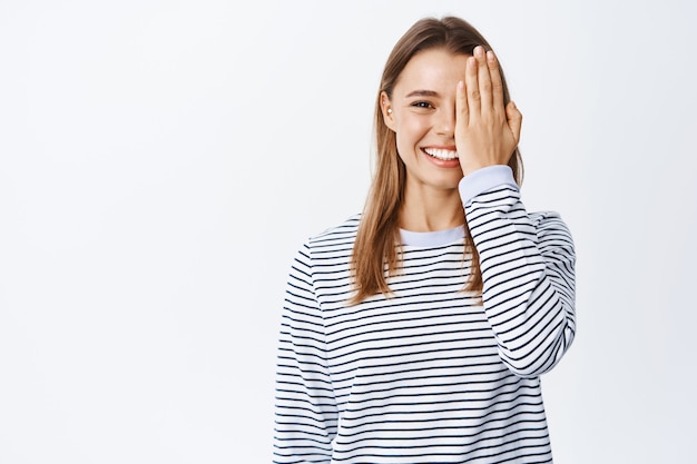 Foto gratuita ragazza bionda allegra con un sorriso bianco, che copre metà del viso con la mano e fissa davanti, prima dell'effetto, muro bianco