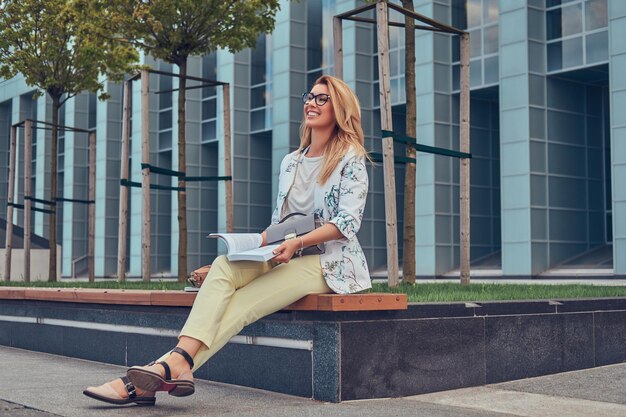 Cheerful blonde female in modern clothes, studying with a book, sitting on a bench in the park against a skyscraper.