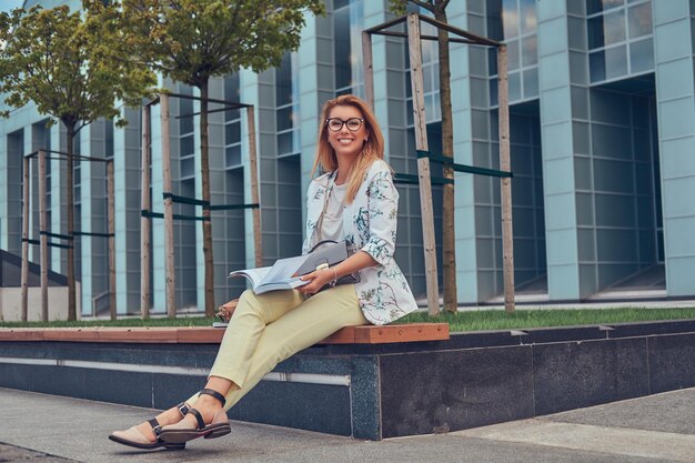 Cheerful blonde female in modern clothes, studying with a book, sitting on a bench in the park against a skyscraper.