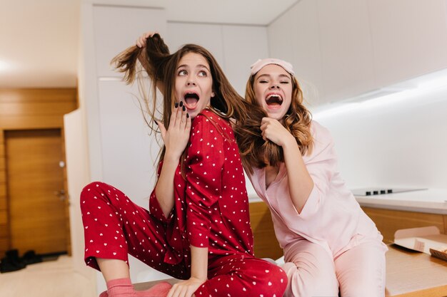 Cheerful blonde girl in sleepmask playing with friend's hair and laughing. Indoor portrait of carefree caucasian sisters fooling around before breakfast.