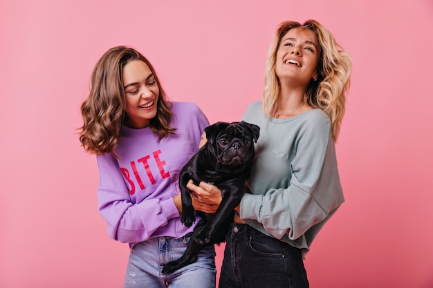 Cheerful blonde girl with happy face expression holding black funny puppy. Indoor portrait of brunette female model looking with smile at her pet.