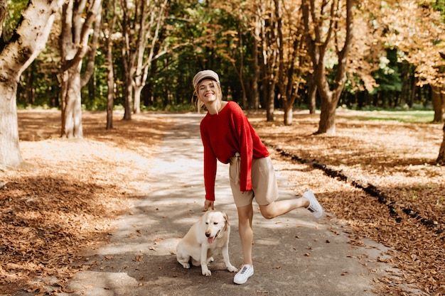 세련 된 빨간 스웨터와 베이지 색 반바지에 밝은 금발 그녀의 강아지와 함께 아름 다운가 공원에서 재미.