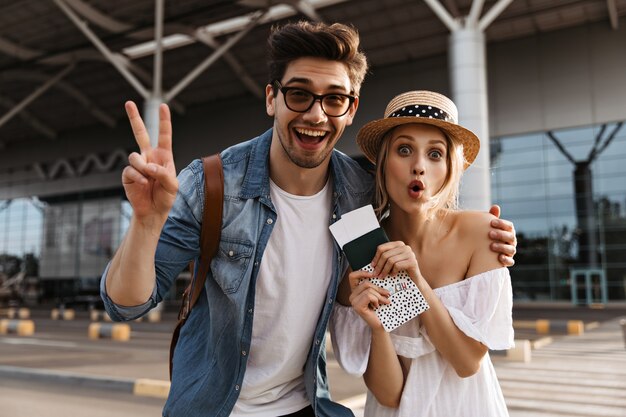 Cheerful blonde woman in hat makes funny face, holds passport and tickets