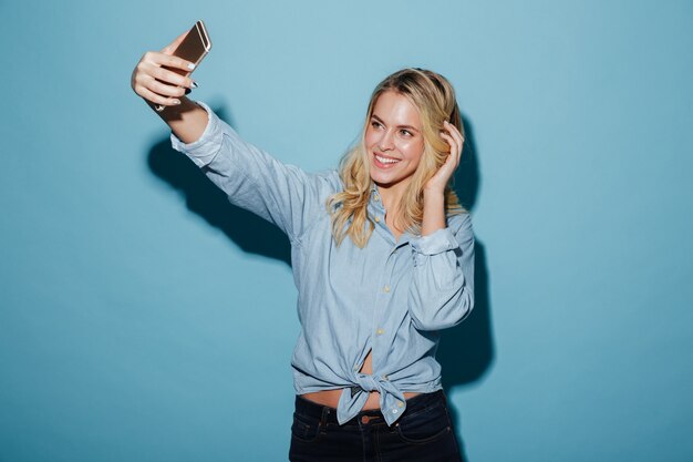 Cheerful blonde woman in shirt making selfie on smartphone