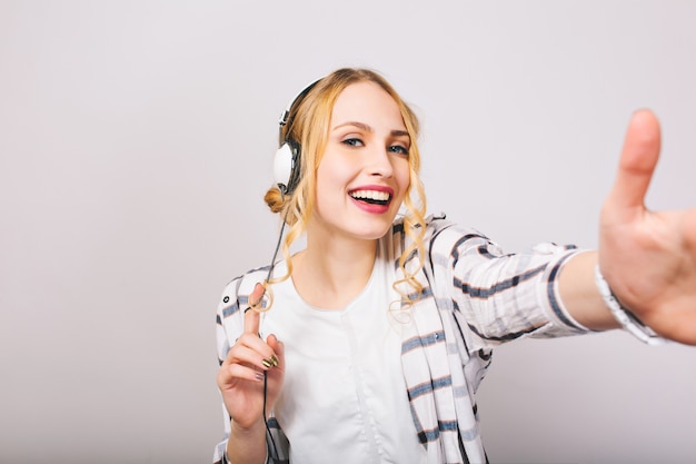 Donna bionda allegra in vestito alla moda bianco che posa nella risata delle grandi cuffie. ragazza graziosa riccia che indossa la camicetta a righe ascoltando musica in nuovi auricolari e divertendosi, ballando