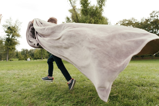 Cheerful boy running with a flying blanket in the park