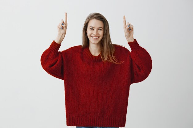 Cheerful bright girlfriend shining from happiness, seeing breathtaking great present. Attractive joyful european model in trendy loose sweater, raising index fingers high, pointing up, smiling broadly