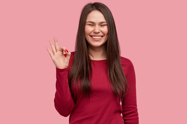 Cheerful brunette Asian lady makes okay gesture with hand, smiles broadly, has freckled face, demonstrates her agreement, wears red clothes