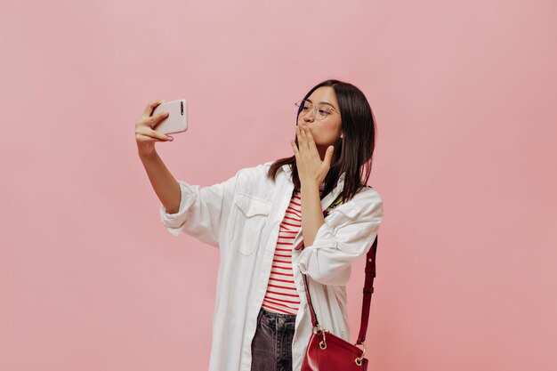 Cheerful brunette Asian woman in oversized white shirt and striped tshirt holds red handbag blows kiss and takes selfie on pink background