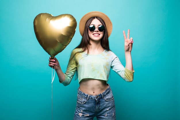 Cheerful brunette beautiful lady in dress holding air golden balloon like heart and showing peace gesture isolated