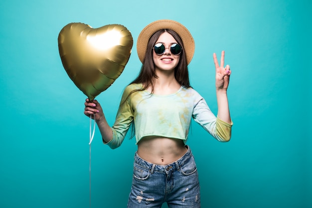 Cheerful brunette beautiful lady in dress holding air golden balloon like heart and showing peace gesture isolated