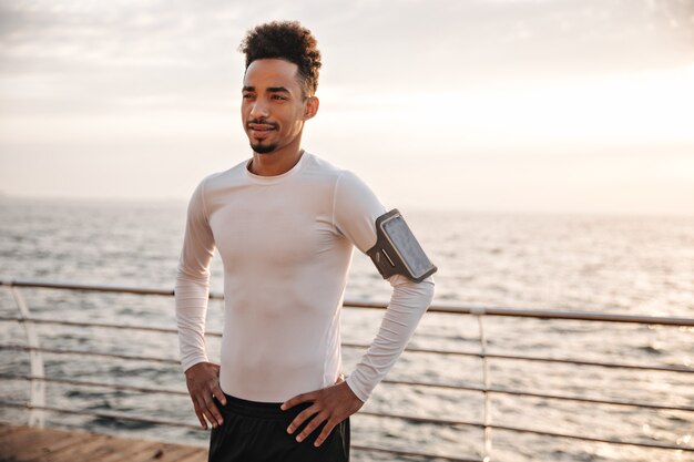 Cheerful brunette curly dark-skinned man in white t-shirt and black sport shorts stands near sea and looks into distance