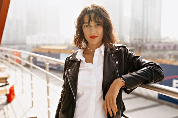 Cheerful brunette curly woman with red lips wearing white shirt and dark jacket is posing on blur city background on great mood outdoors