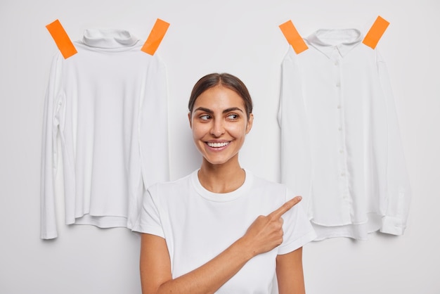 Cheerful brunette European woman points away with index finger demonstrates something advertises clothes dressed in casual t shirt poses against white background two shirts plastered behind