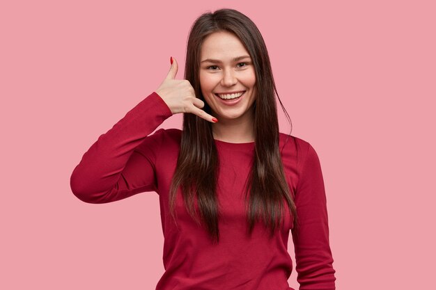 陽気なブルネットのそばかすのある女性は、電話のジェスチャーをし、広い笑顔を持ち、白い歯を見せ、カジュアルな赤いセーターを着て、彼女に電話するように頼みます