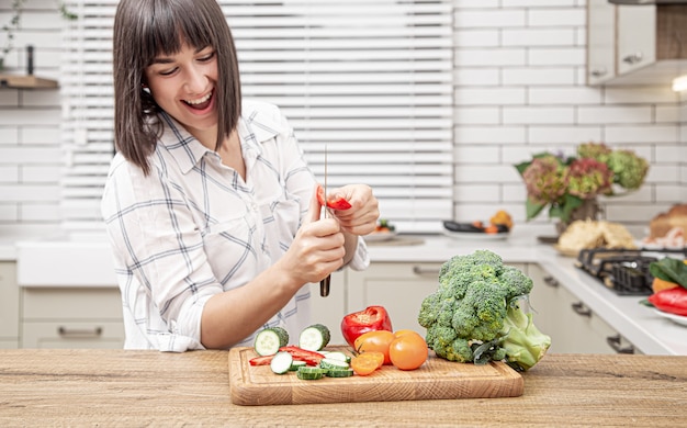 Cheerful brunette girl cuts vegetables on salad on the background of modern kitchen interior.