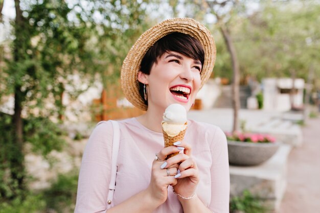 幸せな笑顔で目をそらしているおいしいアイスクリームを保持している陽気なブルネットの女の子