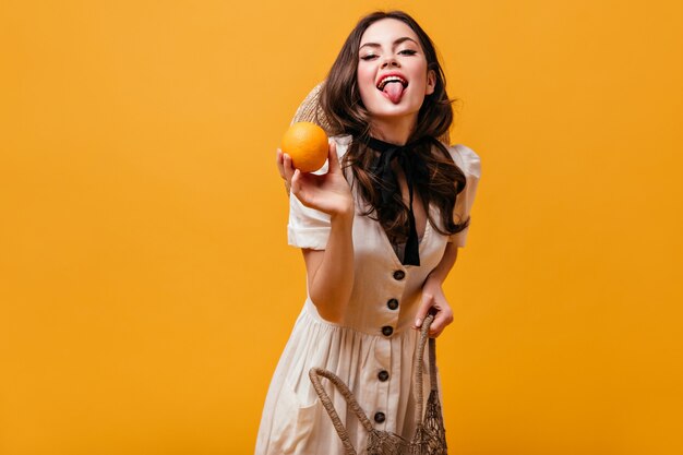Cheerful brunette girl holds shopping bag, orange and shows her tongue on isolated background.