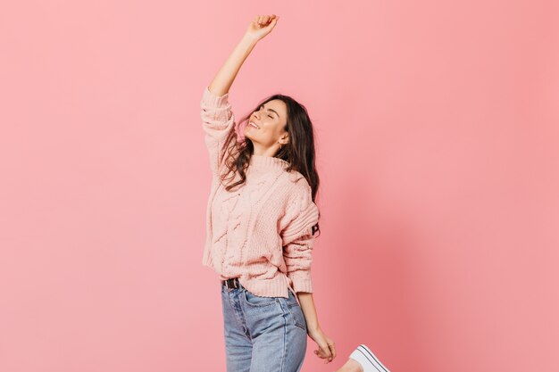 Cheerful brunette girl in knitted sweater and blue jeans raises her hand up and smiles on pink background.