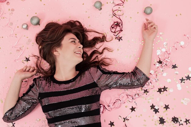 Cheerful brunette girl lies on a pink background among confetti and streamer, the concept of celebrating christmas and new year, festive party.