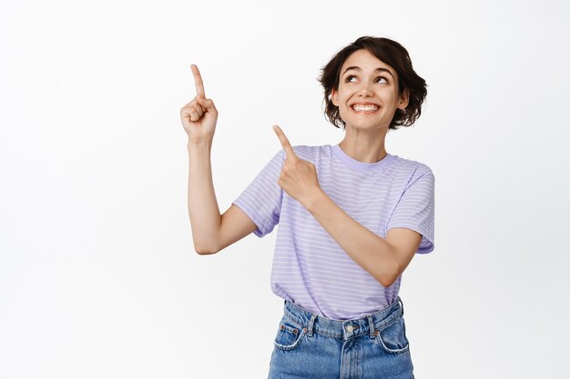 Cheerful brunette girl looks dreamy and happy, pointing fingers at upper left corner standing in t-shirt and jeans on white.