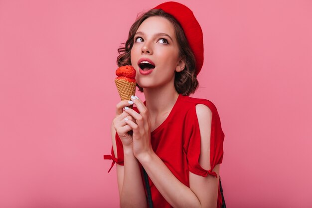 Cheerful brunette girl in red t-shirt posing with ice cream. Indoor photo of winsome lady in beret eating dessert.