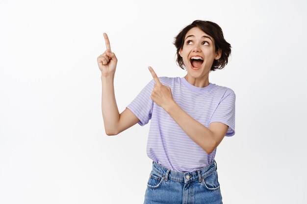 Cheerful brunette girl smiling, laughing and looking excited at info, logo banner, pointing fingers at advertisement upper left corner, empty space, white background