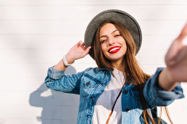 Cheerful brunette girl in white wristwatch and gray hat posing in front of wooden wall touching camera.