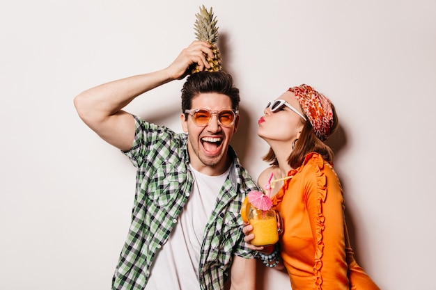 Cheerful brunette man in bright sunglasses holds pineapple on his head while his girlfriend kisses him on cheek.