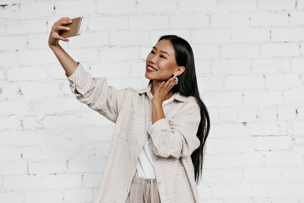 Cheerful brunette woman in beige cardigan smiles sincerely on white brick wall background Tanned lady in good mood takes selfie