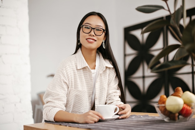 Free photo cheerful brunette woman in eyeglasses, massive earrings and beige jacket smiles and holds cup of coffee