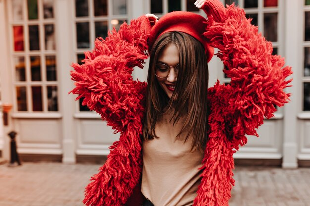 Cheerful brunette woman in glasses is dancing against windows in white frames. woman in beige t-shirt, red beret and coat is having fun on street.