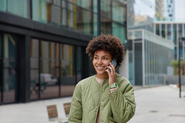 陽気なブルネットの女性は友人と電話で会話をしていますカジュアルなジャケットを着て都会の背景に対して屋外を散歩します幸せな表情を持っています人々現代技術自由時間の概念