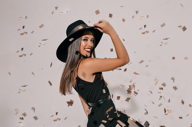 Cheerful brunette woman holding her hat and jumping over isolated wall. Carefree girl enjoying confetti around and have a good mood at party