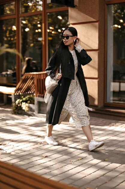 미디 폴카 도트 드레스와 검은색 세련된 트렌치 차림의 쾌활한 브루네트 여성이 바깥으로 이동합니다. 선글라스를 쓴 매력적인 여성이 거리를 걷습니다.