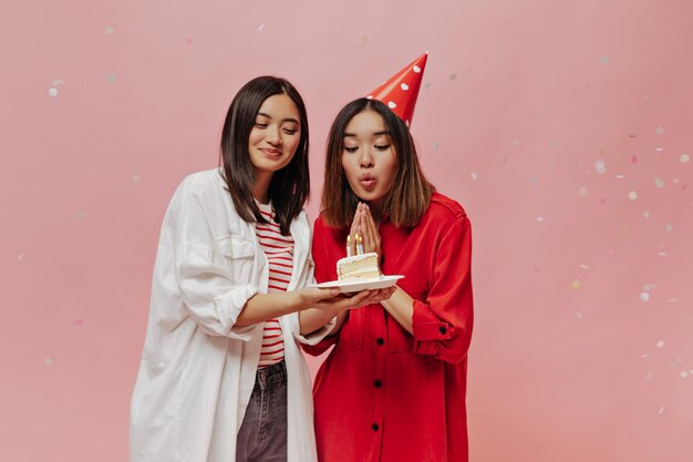 Веселая брюнетка в огромной рубашке улыбается и дарит кусок праздничного торта своей подруге Азиатская девушка в красной лепешке задувает свечи и празднует день рождения