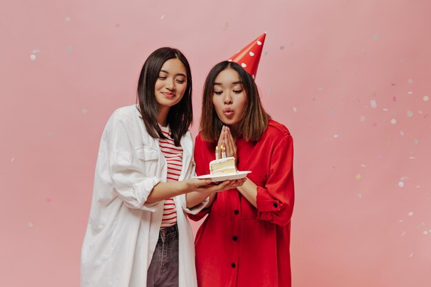 Cheerful brunette woman in oversized shirt smiles and presents piece of birthday cake to her friend Asian girl in red patty hat blows out candles and celebrates bday