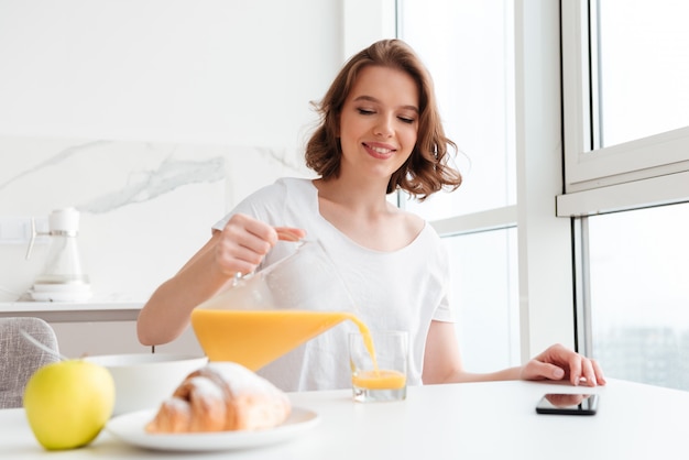 陽気なブルネットの女性が座って、キッチンで朝食をとりながらグラスにジュースを注ぐ