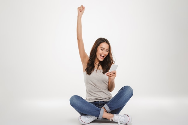 Cheerful brunette woman sitting on the floor and rejoices while using smartphone over gray