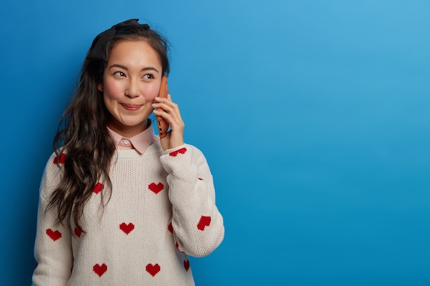 Cheerful brunette woman smiles happily, has telephone conversation, holds smartphone near ear, wears jumper, enjoys nice talk, isolated on blue wall, copy space aside for your promotion