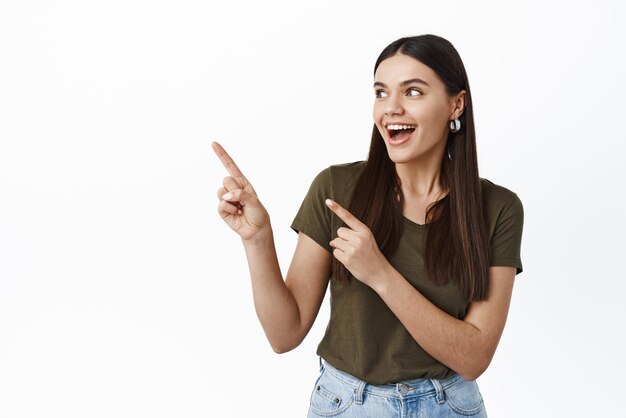 Cheerful brunette woman in tshirt pointing fingers left and smiling looking at copy space logo showing banner with product standing against white background