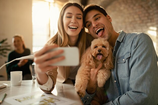 犬と一緒に楽しんで、オフィスで携帯電話で自分撮りをしている陽気なビジネスカップル