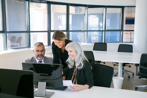 Cheerful business group watching presentation and laughing. Managers sitting at workplace together, looking at computer monitor and laughing. Business communication or teamwork concept