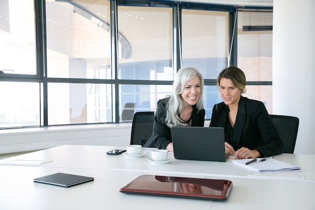 Cheerful business ladies looking at laptop display, talking and smiling while sitting at table with cups of coffee in office. Teamwork and communication concept