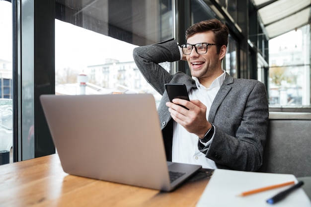 Cheerful businessman in eyeglasses sitting by the table in cafe with laptop computer and smartphone while holding head and looking away