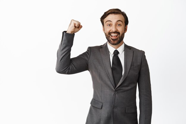 Cheerful businessman rooting for team raising clench fist in support smiling and looking hopeful at camera encourage team standing over white background