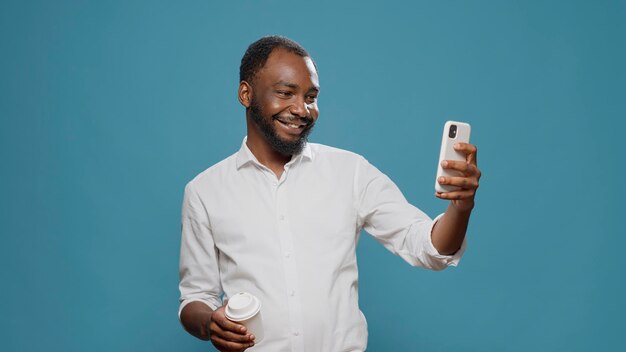 カメラのコーヒーカップでポーズをとって、携帯電話で写真を撮る陽気なビジネスマン。スマートフォンを使ってコーヒーブレイクで写真を撮り、最新のテクノロジーを楽しんでいる幸せなフリーランサー。