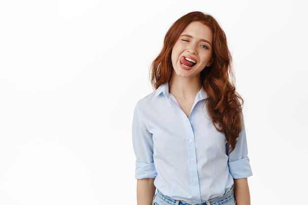 Cheerful candid redhead girl, smiling and showing tongue, winking at you carefree, standing relaxed and joyful against white background. Copy space