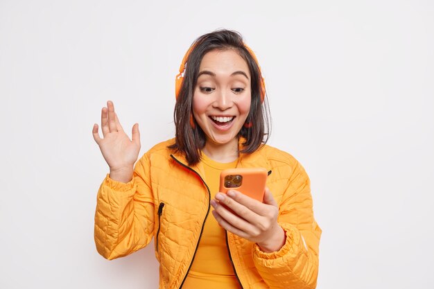 Cheerful carefree Asian teenage girl holds smartphone waves hand makes video call picks song on internet music platform listens favorite podcast uses wireless headphones dressed in orange jacket