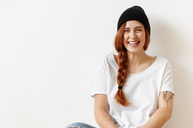 Cheerful carefree cute hipster girl with tattoo sitting on floor at white wall